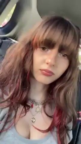 A woman with brown hair and bangs, wearing a gray shirt and necklaces, is sitting in the passenger seat of a car. She looks at the camera and touches her hair. The car interior is visible. The background includes other cars and trees.