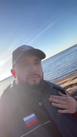The video shows a medium shot of a man, from the chest up, taking a selfie near a body of water, with the sun shining directly behind him. He has short brown hair, a beard, and is wearing a dark blue jacket and a black baseball cap with a logo on it.  A Russian flag patch is visible on the left side of his jacket. His left hand is placed over his chest. In the background, there is water with a sandy shore, and a ship or industrial structure is visible in the distance.  The sky is blue with some thin clouds and contrails.