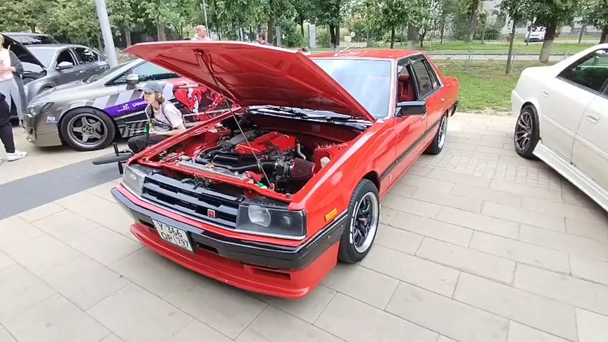 The video begins with a close-up of a red car with its hood open, revealing a meticulously clean engine bay. The engine itself is predominantly red, with black hoses and silver components. A red air filter is visible on the right side of the engine. In the background, another car, gray and white with racing stripes, is parked to the left. A person wearing a white t-shirt and a baseball cap is sitting on a bicycle next to the gray car, looking at the engine. To the right of the red car, a portion of a white car is visible. The scene appears to be outdoors, possibly at a car show or gathering, with trees and buildings in the distance. The camera zooms in slightly on the engine at the beginning of the clip.