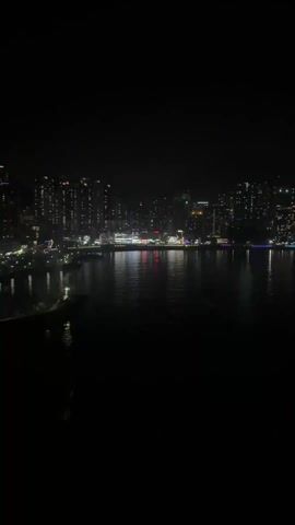 At night, the camera faces a skyline across the water from an unknown location. The lights of many buildings illuminate the cityscape, their reflection shining on the dark water. Occasional colorful light displays highlight some buildings. A cable car system runs across the water in front of the camera, with cars displaying bright green lights.