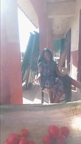 A woman is sitting on a chair outside in front of what appears to be a building. She is eating a piece of fruit, and she looks up to smile at the camera. She is wearing a patterned short-sleeved dress and has very long black braids. Leaning against the wall next to her is a stack of metal sheets. In the foreground of the shot, we see a display of tomatoes. It is daytime and sunny.