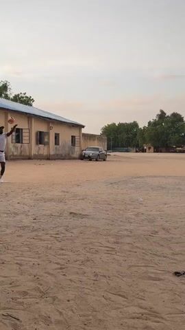 The video captures a volleyball game taking place outdoors on a sandy court. A volleyball net is visible in the center of the frame, separating the two sides of the court. Several individuals, all dressed in white shirts and shorts, are participating in the game. Some players are actively engaged in hitting and passing the ball, while others appear to be observing or waiting for their turn. A group of people are seated on chairs along the side of the court, seemingly watching the game. Trees and buildings are visible in the background, under a bright sky. Litter is scattered on the sand, including small white objects which may be pieces of paper or plastic.