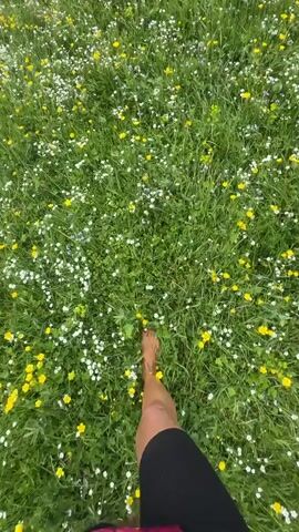 A first-person perspective video captures a person walking barefoot through a vibrant field of grass dotted with numerous small white and yellow wildflowers. The dense green grass is abundant with these blossoms, creating a rich, textured ground cover. The camera angle looks down at the person's legs and feet as they move forward, with their bare feet appearing to sink slightly into the soft earth and brush against the flowers with each step. The person is wearing dark athletic shorts.

The scene is bathed in natural light, suggesting it is daytime, likely midday given the brightness. The overall impression is one of peaceful immersion in nature, evoking feelings of freedom, tranquility, and connection with the natural environment. The abundance of flowers and the lush greenery indicate a temperate season, possibly late spring or summer. No text is visible in the video. The location context is Ketrisi, Georgia.