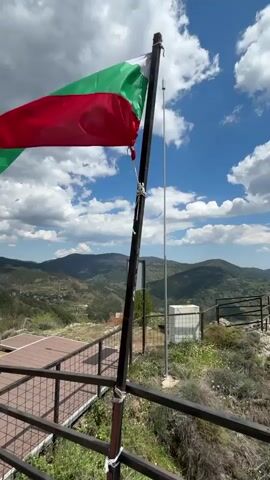 The video shows the Bulgarian flag blowing in the wind, with a scenic mountain landscape in the background. The camera pans out to reveal the landscape and a partially cloudy sky. The flag is attached to a black pole with ropes, and there's a silver pole next to it. The landscape consists of rolling green hills and valleys, stretching out to the horizon. The foreground includes a wooden deck, a black metal railing, and rocky terrain with some vegetation. The sky is blue with fluffy white clouds.