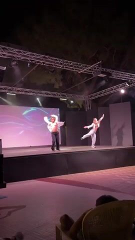 Two young women are performing on an outdoor stage at night. The stage is illuminated by spotlights and has a large screen displaying abstract, colorful graphics behind the performers. Both women are wearing loose-fitting pants and loose white tops. The woman on the left has her hair pulled back in a ponytail and is wearing dark pants. The woman on the right has long, blonde hair and is wearing light-colored pants and a white jacket over a dark, possibly sequined, top. They appear to be dancing and interacting with each other. In the foreground, a person is seated in a chair, facing away from the camera and the stage. The background is dark with trees visible overhead.