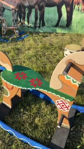 The video pans across a wooden rocking horse outdoors. The rocking horse is painted brown with a tan mane and tail, and features colorful painted designs on its saddle and body, including red flowers and geometric patterns in green, orange, and white. The rocking horse has black handles and a blue rocker base decorated with white wavy lines. In the background, there is a blurred image of horses grazing in a field, and a bale of hay. The ground is covered in green grass.