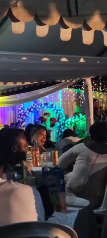 The video shows a gathering of people at what appears to be a formal event, possibly a wedding reception, taking place under a tent adorned with white fabric, string lights, and floral decorations. The floral arrangements consist of white flowers with some red roses. The lighting shifts between green and purple. People are seated at tables with drinks and snacks. Some individuals are turned towards the stage area where a person is standing.  The focus of the video is on the people at the tables.