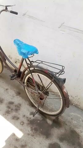 The video shows a brown bicycle with a blue plastic cover over the seat, resting against a white wall. The bike has a black basket attached to the front handlebars and a rack above the back wheel. There is a kickstand deployed. The bicycle appears to be old and has some rust. The ground is a textured concrete surface with some stains. The camera moves slowly around the bicycle.