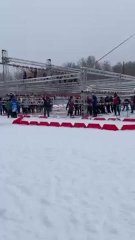 The video shows a winter obstacle course event. The ground is covered in snow, and several people are wearing winter jackets and hats, indicating a cold environment. Overhead, a large, multi-level metal structure is visible, with participants attempting to traverse obstacles on it. A crowd of people is gathered around the base of the structure, presumably observing or waiting for their turn. There are red mats or barriers on the ground. The sky is overcast, suggesting a dull or snowy day.