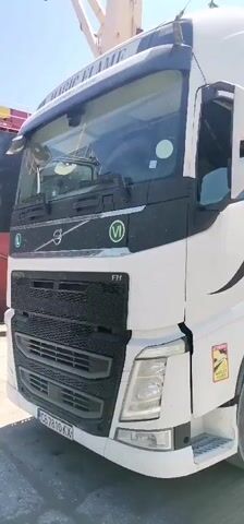 A white Volvo truck with a trailer is parked on a paved surface. The trailer is a tipper trailer and is raised at an angle, with its contents being emptied. Large, gray, rocky material is falling from the trailer onto a pile of similar material. In the background, a large ship is docked, and a tall, orange and green crane is operating, likely loading or unloading cargo from the ship. The scene appears to be a port or industrial area, with a clear blue sky overhead.