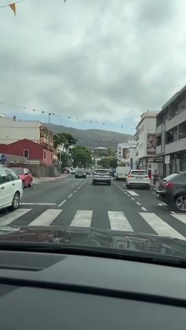 A view from inside a car traveling down a street lined with buildings. The sky is overcast and colorful triangular flags are strung across the street. There are several cars on the road, including a red car on the left, a black car in the center, and a white SUV on the right. A pedestrian is walking on the sidewalk on the right side of the street. In the distance, there are mountains.