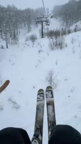 The video shows a first-person perspective from a ski lift ascending a snowy mountain. Two skis, each with "S3" printed on them, are visible in the foreground, along with the skier's legs clad in black ski pants. To the left, the edge of another ski is partially visible, indicating the presence of another skier in the lift. The lift carries the skier up a slope covered in snow, with scattered small trees and shrubs. A ski lift tower is visible further up the hill. The weather appears overcast.