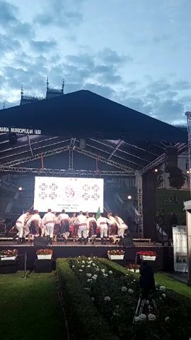 The video shows a group of people in traditional clothing dancing on a stage. There is a large screen behind them displaying an image. In front of the stage, there is a garden with flowers. The sky is cloudy.