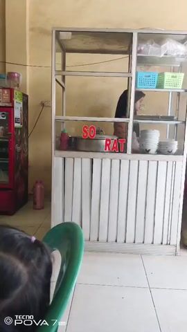 The video shows a food stall with a glass display case. There are shelves inside the case, some with blue and green baskets, and bowls stacked on another shelf. A sign with red lettering that says "SO RAT" is visible on the display case. A person wearing a headscarf is partially visible behind the counter. In the foreground, the back of a person's head with dark hair is visible, with a green chair in front of them. To the left of the stall, a red refrigerator is visible, and to the right, a yellow gas tank stands on the floor. The stall appears to be indoors, with tiled flooring and yellow walls.