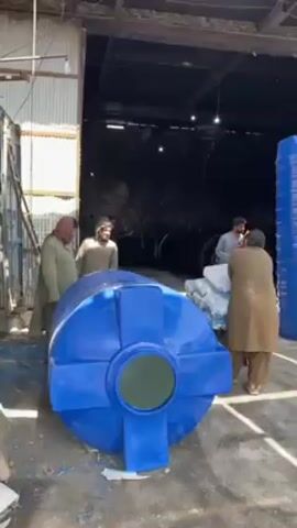 The video shows a warehouse scene where several men are working. A large blue plastic water tank lies horizontally in the foreground. Four men are visible, dressed in traditional attire, moving around the tank and a stack of packaged materials. In the background, the warehouse interior appears dim, with glimpses of what seem to be larger tanks or equipment. The warehouse has a corrugated metal roof and walls, with natural light entering from a large opening. The ground is concrete, showing some signs of wear and debris.