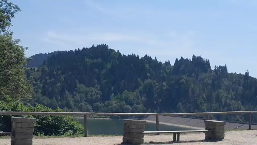 The video shows a wide shot of a hilly, tree-covered landscape with a body of water visible in the background. In the foreground, there is a wooden fence and two stone pillars. A bench is also visible behind the fence. The sky is clear and blue.