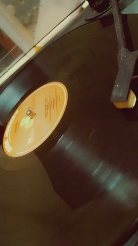 The video shows a close-up of a vinyl record playing on a turntable. The record is black with an orange label in the center. The label has text and graphics on it, but they are not clear enough to read. The turntable's needle is visible and appears to be in contact with the record. The background is dark and slightly out of focus. The video is shaky, suggesting it was filmed handheld.
