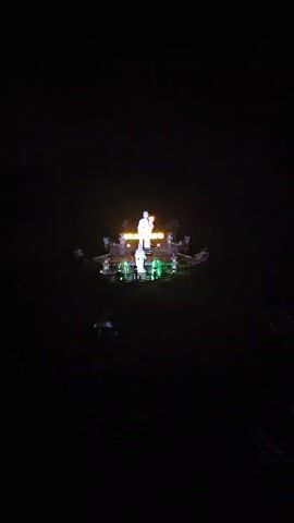The scene is a nighttime view of a brightly illuminated Buddhist temple against a dark sky. The temple features a traditional Asian roof design with elaborate ornamentation. Dominating the structure are two white statues, the taller one standing atop a slightly elevated platform. The temple is adorned with green lights along its base, and an illuminated sign is visible above the statues. The temple is surrounded by the dark silhouettes of trees. The camera slowly zooms in on the temple, providing a closer view of the statues and architectural details. The video is static and lacks audio.