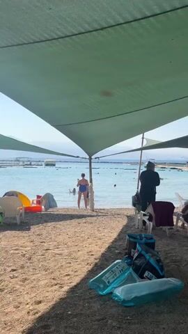 The video shows a beach scene under a large green canopy providing shade. In the foreground, several white plastic chairs are scattered on the sand. Some inflatable water toys and bags are on the ground in front of the chairs. In the midground, several people are visible. To the left, a woman in a blue swimsuit stands near the water's edge. Further back, a person wearing a black jacket and hat stands near a chair. To the right, two women are talking. Other people are seated in chairs or standing around the beach. In the background, the blue ocean stretches to the horizon, with some boats visible in the distance. Trees and foliage can be seen beyond the beach. The lighting suggests it is daytime, likely a sunny day.