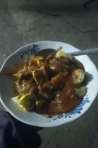 The video shows a close-up view of a plate of food. The food is served in a white bowl with blue floral accents. A spoon is resting on the edge of the bowl. The dish appears to be a savory meal with chunks of what looks like tofu and possibly some kind of meat or dumpling, all covered in a thick, reddish-brown sauce that resembles peanut sauce. The background is dark and appears to be an outdoor setting with a rough, concrete-like surface. The lighting is dim, suggesting it might be nighttime or an indoor location with low light. The camera is held steady, focusing on the food. There is no visible interaction or movement other than the slight jostling of the bowl and spoon.