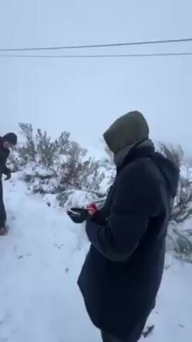 The video shows a snowy scene with three people, two of whom are visible from the waist up. One person is wearing a long, dark-colored coat with a hood, and black gloves. The other person has a dark jacket. They are walking on snow-covered ground. The road has snow on each side, and at least three cars are visible, driving along the road. The sky is overcast.