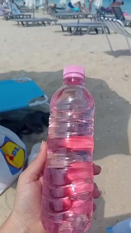 The video begins with a close-up of a hand holding a plastic water bottle filled with pink liquid and ice, at a sandy beach. In the background, beach chairs and umbrellas are visible. The camera then pans to show the beach environment. A woman is sitting under a large umbrella, wearing a black bikini. A man is relaxing on a blue lounge chair, looking at his phone. In the distance, people are swimming and walking in the sea, under a clear blue sky with scattered clouds. The scene captures a typical day at a beach resort.