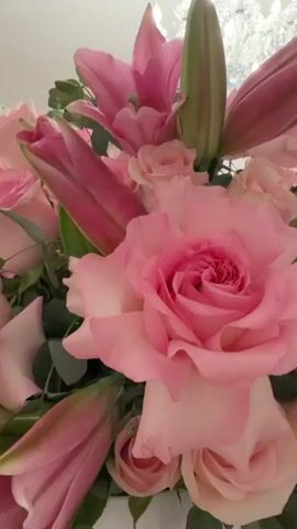 A close-up shot of a large floral arrangement of pink roses and lilies is shown against a background featuring a crystal chandelier and artwork on a wall. The camera slowly pans upwards, revealing more of the chandelier and the room's decor. The lighting is bright, highlighting the vibrant colors of the flowers and the sparkle of the chandelier.