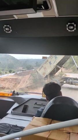 From the perspective of someone seated in a vehicle, a white dump truck is actively depositing gravel onto a dirt road. The truck's bed is raised, and a man in a brown shirt is standing nearby, possibly directing the dumping process.  The truck bears the name "M.K.R MAHADEV" in large blue letters on the front. The road is unpaved and appears to be under construction, with hills visible in the background. The vehicle containing the camera operator appears to be waiting for the dump truck to complete its task.