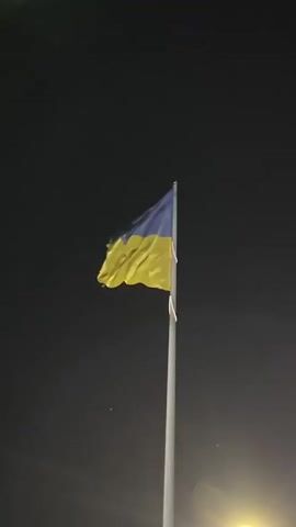 A Ukrainian flag waves on a flagpole against a dark sky. The flag is positioned slightly to the right of the center of the frame. The sky is mostly black, with some faint, blurry lights visible at the bottom of the frame, suggesting artificial lighting. The flagpole is a light gray color and extends from the bottom of the frame upwards, holding the flag. The flag itself is horizontally divided into two equal bands of color: blue on the top and yellow on the bottom. The flag is captured in motion, with visible ripples and folds. The overall lighting of the scene is low, indicating it is likely nighttime or dusk.