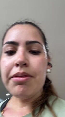 A young woman with dark hair pulled back is shown in a close-up, low-angle shot, looking directly into the camera. Her face and neck are visibly sweaty, with beads of sweat on her forehead, cheeks, and chin, indicating recent physical exertion. She has defined eyebrows and long eyelashes. She is wearing a light green t-shirt, a delicate gold chain necklace with small beads, and a white pearl earring that appears to be integrated with a black earbud or wire in her right ear.

At the 0:01 mark, she lifts her right hand to push her hair back from her forehead, her expression shifting slightly. She exhales audibly and her mouth is slightly open, suggesting heavy breathing. She then looks upwards and slightly to the right, her eyes focused, with a determined or tired expression. The background is a plain, light-colored ceiling with some recessed lighting fixtures visible. Her skin is flushed from exercise. She continues to look upwards and to the side, her breath still heavy, until the video ends.