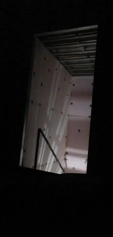 The video shows an unfinished interior space, possibly a room or a closet, viewed from an elevated angle looking down. The walls are covered with pink insulation boards, some of which have dark circular fasteners or marks. Metal framing is visible along the ceiling, suggesting that a finished ceiling will be installed. A black railing or structural element is present in the foreground, partially obscuring the view of the room. The lighting is dim, with some shadows cast on the walls, creating a somewhat eerie atmosphere. The overall impression is of a construction or renovation site that is in its early stages.