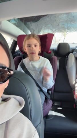 A woman with dark hair and sunglasses is driving a car while recording a video from the driver’s seat. Her daughter, a young girl with light brown hair, is in a car seat in the back, singing along to music. The car has a sunroof. The mother gestures with her hand as she records. The daughter gestures along to the music as well. The car has black interior with a burgundy-colored headrest on the car seat in the back.