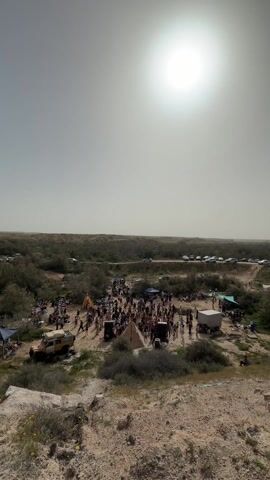 From an elevated perspective, the video shows a gathering of people in a desert-like environment. The scene is bathed in bright sunlight under a hazy sky. The group of people, numbering in the dozens, are gathered in an open area with sparse vegetation and light-colored sand.  Several vehicles are parked nearby, including a yellow jeep to the left and a row of cars along a dirt road in the background.  There are a few tents and small shelters erected, including a triangular tent in the center of the gathering.  Speakers and sound equipment are positioned around the tents, suggesting a party or event with music. The background features low rolling hills and scattered greenery, providing a sense of vastness to the location. The overall atmosphere suggests an outdoor gathering, possibly a festival or party, in a remote or natural setting.