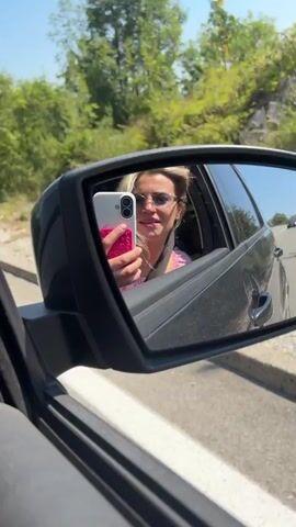 The video shows a woman with blonde hair and sunglasses in the passenger seat of a car, holding up a phone and taking a selfie in the side-view mirror. The background is blurred trees and a road, suggesting the car is in motion. The woman is wearing a pink top and appears to be smiling and talking to the camera.