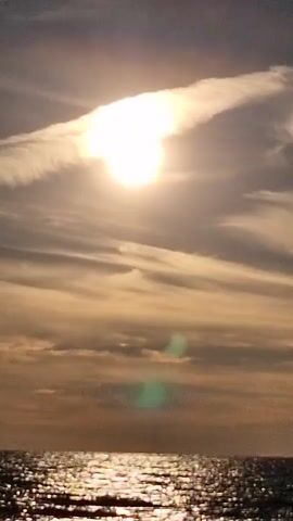 A vertical video shows a bright sun partially obscured by clouds over the ocean. The sun is near the top of the frame, emitting a strong glare. Wispy clouds stretch across the sky, with some darker clouds lower down. The ocean surface reflects the sunlight, creating a shimmering effect. The overall scene is calm and serene.