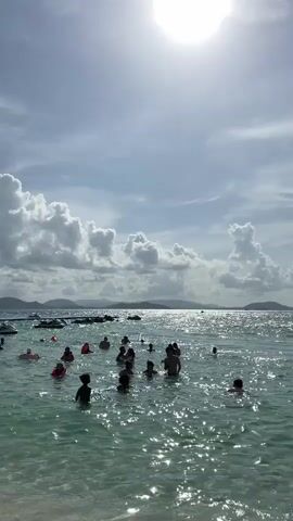 A group of people are swimming in clear, shallow ocean water under a bright, sunny sky with scattered clouds. Some of the people are wearing life vests. There are jet skis and other small boats visible in the distance near the shore. The water is calm with some light waves and sunlight glinting off the surface. The scene appears to be a popular beach or swimming area.