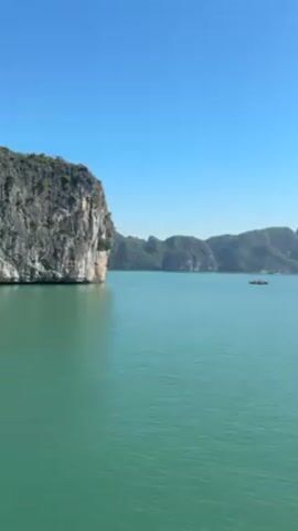 The video shows a wide shot of Halong Bay. The water is a turquoise-green color and the sky is clear blue. There are several rocky islands with greenery on top, dotting the horizon. A small boat appears in the distance. The video appears to be taken from a boat, panning slowly to the right.