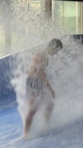 A person is surfing on a wave machine. The person is wearing swim trunks and is being splashed by water. The background is a wall with windows. Another person, a girl, is visible in the foreground, also in the water.