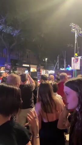 A group of people are gathered outdoors at night. They are looking towards a large screen that is displaying an image. There is a DJ booth with a person standing behind it. A tall pole with lights is visible in the background, along with a red banner that says "DUCATI". The people are casually dressed and some are holding up their phones, likely to take pictures or videos. The atmosphere appears to be festive, possibly a street party or an outdoor event.