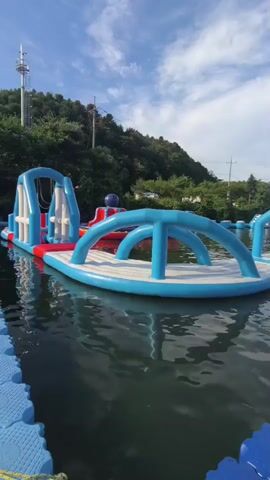 The video shows a floating water park on a sunny day. There are several inflatable structures in the water, including slides and obstacle courses. A blue and white floating walkway leads to a tent structure in the background. The water appears calm, and the sky is blue with some clouds. The overall impression is one of a recreational activity on a lake or calm body of water.