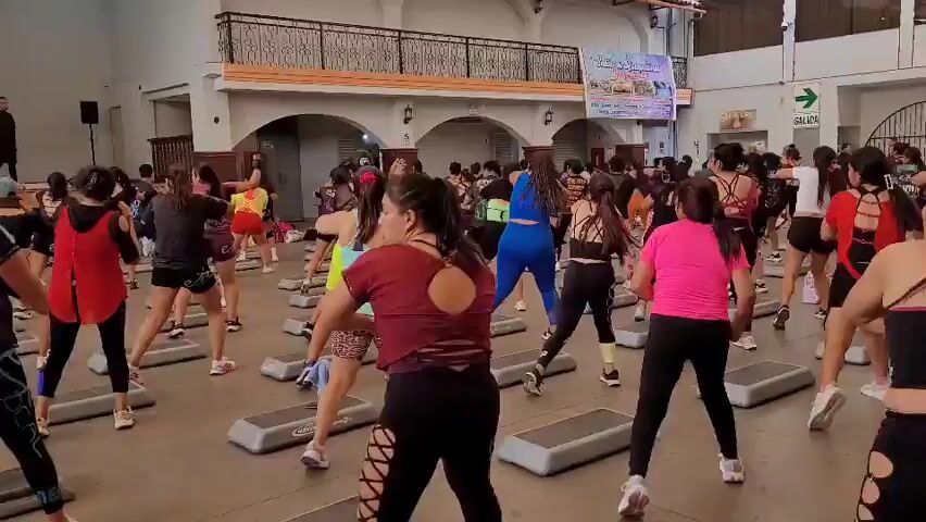 This video captures an aerobics class in a large indoor space with a high ceiling. The floor is concrete, and the walls appear to be painted in light colors with arched windows. In the background, there is a balcony or mezzanine area. Multiple participants, predominantly women, are arranged in rows facing forward, each standing on a grey aerobic step. They are all dressed in athletic attire, including t-shirts, tank tops, shorts, and leggings, and many are wearing athletic shoes. The participants are actively engaged in a choreographed routine, performing various movements such as punches, kicks, and arm gestures. A man in the foreground, wearing glasses and a black t-shirt, is looking to his right and appears to be participating in the class. Another man, dressed in a dark t-shirt, is standing to the far left of the frame, possibly as an instructor or observer. In the background, a banner with text and images is displayed on the wall. The overall atmosphere suggests a fitness event or class session.