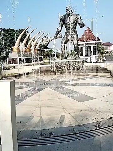 The video shows a statue of a muscular man, possibly a depiction of a character from mythology or folklore, standing in the center of a fountain.  He is holding a large, club-like weapon.  Behind the statue is another sculpture featuring curved, horn-like shapes. Water is spraying from the fountain, creating a spectacle around the statue. A small building with a tiled roof is in the background. The sky is clear and blue. The fountain area appears to be a public space, possibly a park or plaza.