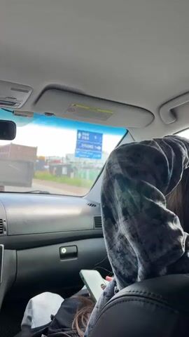 The video shows the interior of a car, looking forward through the windshield. The sun is shining, and the sky is visible. There are two people in the car, with the back of one person's head and shoulder in the foreground, wearing a grey hooded sweatshirt. This person is looking at their phone. Another person is partially visible in the passenger seat, wearing a black jacket and white shirt. Outside the car, there are some buildings, a large truck, and a blue road sign with Korean text and arrows indicating directions. The road sign is the central focus outside the car.