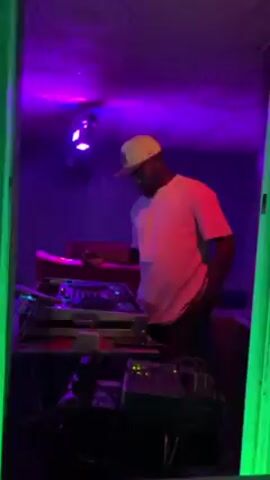 A man wearing a white t-shirt and a baseball cap stands at a DJ console in a dimly lit room. The console has multiple turntables and various audio equipment, with wires and cables visible. The room is illuminated with colorful lighting, including red, green, and blue. The camera angle is from slightly below and to the side, looking up at the DJ. The shot is somewhat obscured by a green post or wall in the foreground. The DJ appears to be adjusting the controls on the console.