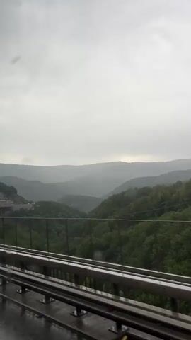 The video shows a view from a moving vehicle on a wet road, looking out over a valley filled with rolling green hills. A metal guardrail is visible in the foreground. The sky is overcast and gray. In the distance, a structure that appears to be a dam or a large building is visible on the side of a hill. The overall impression is of a landscape viewed through a rain-streaked window.