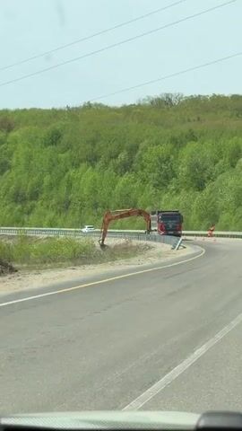 The video shows a red Shacman X3000 dump truck driving along a winding highway curve. The truck is carrying the arm of an excavator in its bed. Orange traffic cones are placed along the right side of the road. On the other side of the guardrail, a green forested area stretches into the distance. The sky is bright, suggesting a sunny day. The view is captured from inside another moving vehicle.
