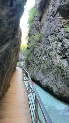 The video shows a person walking on a wooden boardwalk that winds along the side of a narrow gorge. The gorge walls are steep and rocky, with some vegetation growing on them. A turquoise river flows rapidly through the bottom of the gorge. The boardwalk has a metal railing. The person is seen from behind and is wearing dark clothing. The sun is shining from the left side of the frame, casting shadows.