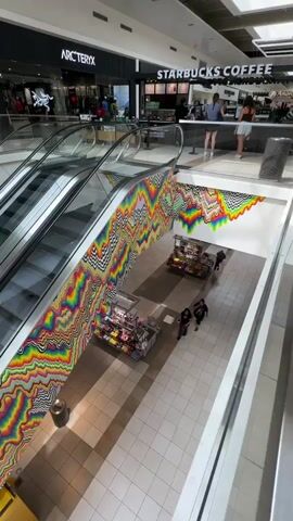 The video shows the interior of a shopping mall. An escalator ascends from the lower level to the upper level. The wall next to the escalator is decorated with a large, colorful, psychedelic mural featuring zig-zag patterns in various rainbow hues. On the upper level, Starbucks Coffee is visible, with several customers standing in line and baristas working. To the left of Starbucks is an Arc'teryx store, and further down the hall are UGG and other retail stores. On the lower level, beneath the Starbucks and Arc'teryx stores, are display tables with merchandise. A few shoppers are seen walking on the lower level. The mall appears well-lit with natural light coming from skylights in the ceiling.