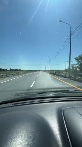 This is a first-person perspective video, presumably from inside a vehicle, looking out through the windshield. The viewpoint is focused on the road ahead, which is a paved highway stretching into the distance under a clear, bright blue sky. The sun is visible in the upper left quadrant of the frame, casting lens flares and creating a bright glare. There are power lines running parallel to the road on the right side, supported by utility poles. The road itself has white lane markings. On either side of the highway, there is a grassy verge, and beyond that, flat, open terrain with some distant trees or bushes. The dashboard of the vehicle is visible in the foreground, occupying the bottom portion of the image. The overall scene depicts a sunny day driving on a highway.