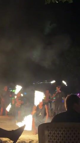 A group of individuals are performing a fire dancing show on a beach at night. One person in the foreground is kneeling and swinging a flaming prop that resembles a fan. Other performers are visible in the background, also wielding flaming objects and tossing them in the air. The area is dimly lit with the flames providing most of the illumination. Smoke is visible in the air, and a crowd of spectators is implied by the presence of seated figures and blurred shapes in the background. The environment appears to be a sandy beach with trees and possibly some structures or lights in the distance.